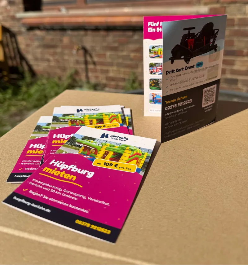Gedruckte Hüpfburg-Flyer auf einem Tisch, dahinter steht ein Flyer aufrecht mit der Rückseite: Kontaktdaten und QR-Code